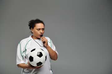 A female referee blows her whistle while holding a soccer ball.