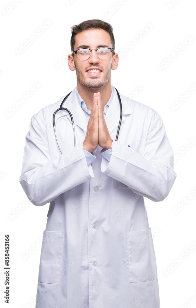 Sticker Handsome young doctor man praying with hands together asking for forgiveness smiling confident.