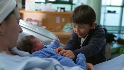 Bonding family moment of sibling interacting with his newborn baby brother at hospital after childbirth while mother watches laid in bed, young boy tickling infant