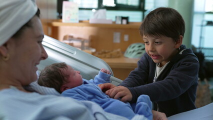 Bonding family moment of sibling interacting with his newborn baby brother at hospital after childbirth while mother watches laid in bed, young boy tickling infant