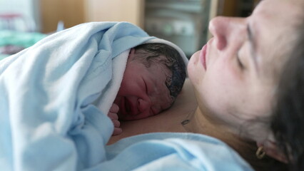 Crying newborn baby resting on mother's chest at hospital clinic right after birth, real life authentic childbirth scene of new infant arriving to this world
