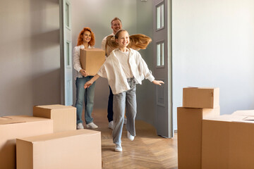Happy child girl running indoors, exploring new big house moving in, parents holding boxes entering own home with kid daughter