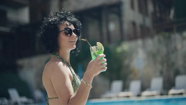 Joyful woman in sunglasses holding a mojito cocktail, dancing by the poolside with a smile, enjoying a sunny vacation day.