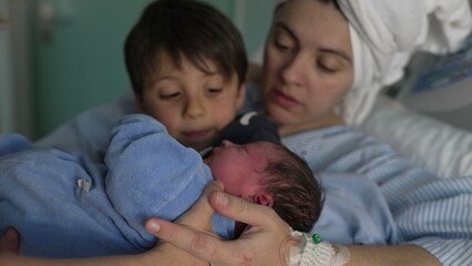 Mother and child welcoming newborn baby in arms at hospital after childbirth. nurse handing little infant to brother's arm next to mom at clinic, siblings bonding for the first time