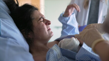 Mother trempling with pain right after giving birth to newborn baby, doctor and nurses surrounding her with care in maternity clinic hospital, hands gently putting infant into chest