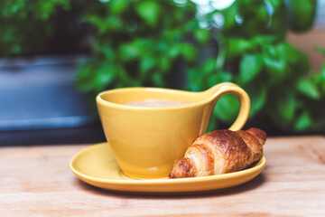 Yellow cup with coffee and crosant on a plate set on a wooden board, breakfast time, French breakfast, coffee break