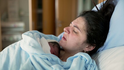 New mother in discomfort after post-partum trauma at hospital clinic holding newborn on chest. woman in pain after childbirth, real life authentic emotion of human struggle
