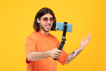 Cheerful middle eastern guy taking photo by smartphone with selfie stick on yellow background.	
