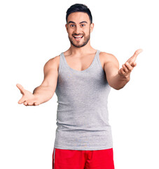 Fototapeta premium Young handsome man wearing swimwear and sleeveless t-shirt smiling cheerful offering hands giving assistance and acceptance.