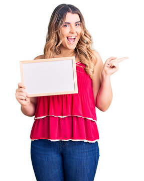 Young caucasian woman holding empty white chalkboard smiling happy pointing with hand and finger to the side