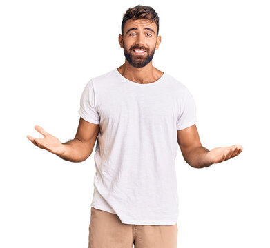 Young hispanic man wearing casual white tshirt clueless and confused expression with arms and hands raised. doubt concept.