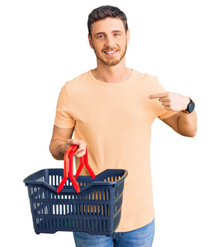 Handsome young man with bear holding supermarket shopping basket pointing finger to one self smiling happy and proud