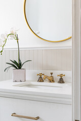 A farmhouse bathroom faucet detail with a white cabinet, gold faucet and hardware, gold circular mirror, and plant sitting on the marble countertop.