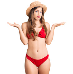 Beautiful caucasian young woman wearing bikini and summer hat clueless and confused expression with arms and hands raised. doubt concept.