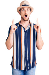 Young handsome caucasian man wearing summer hat amazed and surprised looking up and pointing with fingers and raised arms.