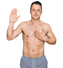 Handsome young man wearing swimwear shirtless swearing with hand on chest and open palm, making a...
