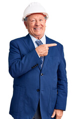 Senior handsome grey-haired man wearing suit and architect hardhat cheerful with a smile of face pointing with hand and finger up to the side with happy and natural expression on face