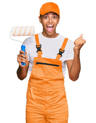 Young handsome african american man wearing cap and painter clothes holding painting roll screaming proud, celebrating victory and success very excited with raised arms