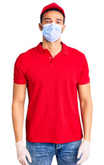 Young handsome hispanic man wearing delivery uniform and medical mask with serious expression on face. simple and natural looking at the camera.