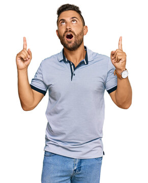 Handsome man with beard wearing casual clothes amazed and surprised looking up and pointing with fingers and raised arms.