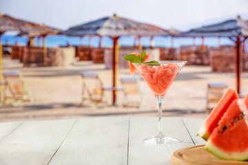 Slices of watermelon and small coctail in glass on white table top and blur background of exotic sandy beach,sun umbrellas and blue sky. Empty copy space for products and food.