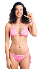 Naklejka premium Young beautiful hispanic woman wearing bikini smiling and confident gesturing with hand doing small size sign with fingers looking and the camera. measure concept.