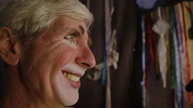 Close-up of face profile of mature male Caucasian circus performer with clowns makeup grimacing and making faces in front of mirror in dressing room