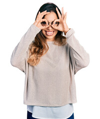 Beautiful hispanic woman wearing casual sweater and glasses doing ok gesture like binoculars sticking tongue out, eyes looking through fingers. crazy expression.