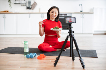 Woman in red activewear sitting on yoga mat eating salad while filming fitness video in modern...
