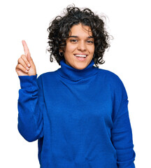 Young hispanic woman with curly hair wearing turtleneck sweater pointing finger up with successful idea. exited and happy. number one.