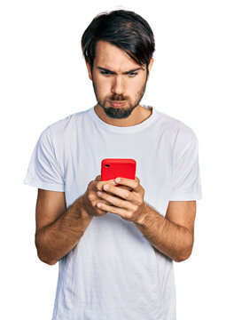 Hispanic man with blue eyes using smartphone typing message puffing cheeks with funny face. mouth inflated with air, catching air.
