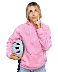 Young caucasian woman holding bike helmet serious face thinking about question with hand on chin, thoughtful about confusing idea