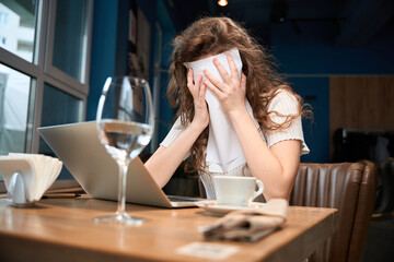 Young woman covered her face with a napkin while sitting and looking at a laptop