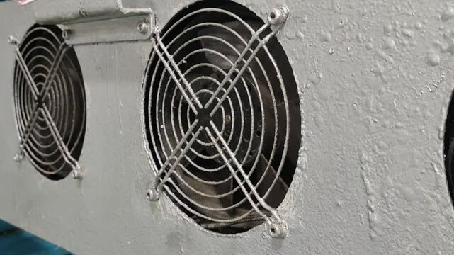 Industrial cooling fans mounted on a metal panel. The fans are circular and have a black metal grille. The panel is painted white and has a weathered appearance