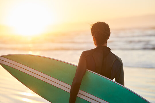 Sunset, surfboard and back of person at ocean for fitness, exercise and wellness in summer. Wetsuit, athlete and challenge or water sports at beach for adventure, cardio and balance in California