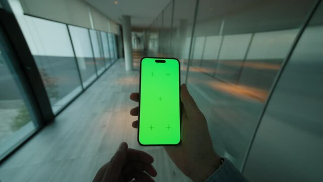 Hand man holding and scrolling vertically phone with green screen. On background modern space with big windows. Typing on smartphone
