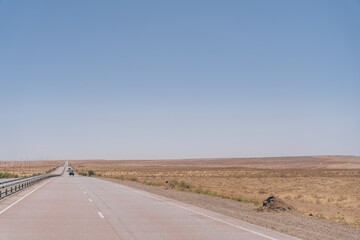 A long, empty road with a blue sky above. The road is lined with tall grass and there is a car driving down the road