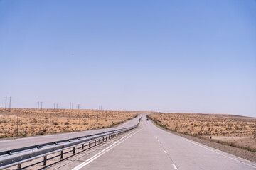 A long, empty road with a blue sky above. The road is lined with tall metal guardrails