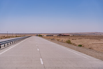 A long, empty road with a few buildings in the distance. The sky is clear and blue