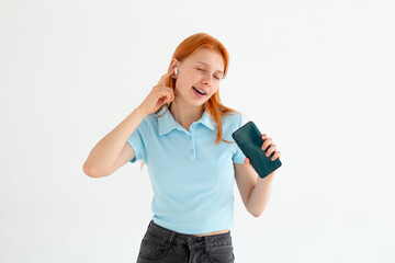 Attractive smiling young redhead woman wearing wireless earphones to listen to music, holding mobile phone