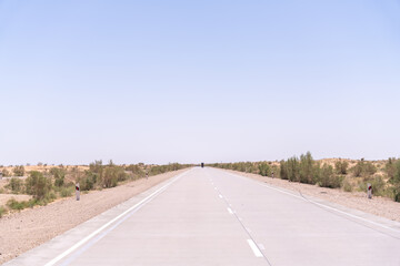A long, empty road with no cars or people. The sky is clear and blue. The road is made of concrete and is very wide