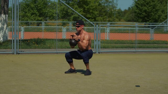 Shirtless young man training on city sports ground and performs squats and bend-overs with kettlebell. Works quadriceps, gluteus maximus, lower back, hamstrings and calves