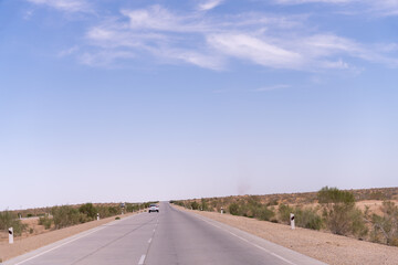 A long, empty road with a car driving down it. The sky is clear and blue. The road is surrounded by a desert landscape