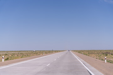 A long, empty road with no cars or people. The sky is clear and blue. The road is made of concrete and has white lines painted on it