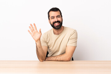Caucasian man with beard in a table saluting with hand with happy expression.