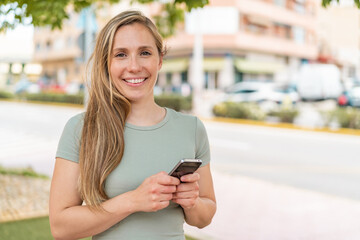 Young blonde woman using mobile phone at outdoors smiling a lot