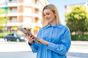 Young pretty blonde woman at outdoors holding a tablet with happy expression