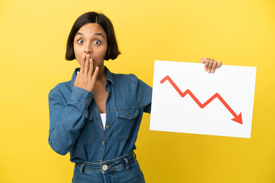 Young mixed race woman isolated on yellow background holding a sign with a decreasing statistics arrow symbol with surprised expression