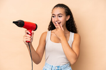 Young woman holding a hairdryer isolated on beige background with surprise and shocked facial...