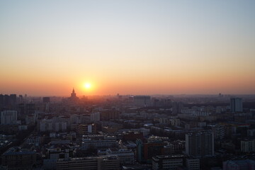 Aerial view of a cityscape at sunset, with the sun descending on the horizon. The silhouette of urban buildings is softly illuminated by the warm, golden light, creating a calm and serene atmosphere. 